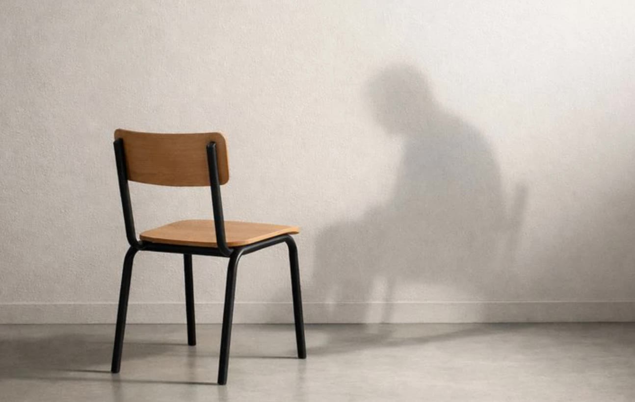 An empty chair against a bare wall; the shadow of a seated man falls where no one sits.